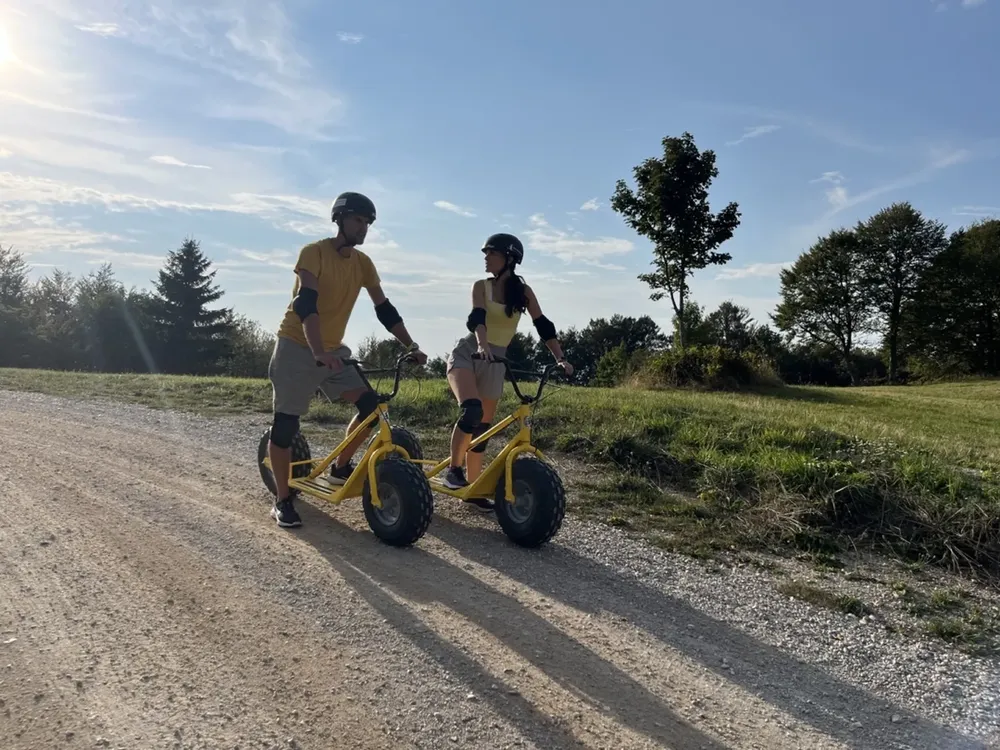 Two people riding mountain scooters downhill from Slivnica on a forest gravel trail with views of the surrounding landscape.