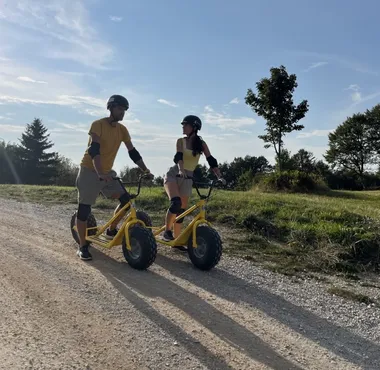 Two people riding mountain scooters downhill from Slivnica on a forest gravel trail with views of the surrounding landscape.