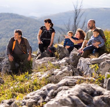 Skupina odraslih in otrok z vodnikom med pohodom počiva na razgledni skalni točki ter občuduje naravo in razglede po okoliških hribih.