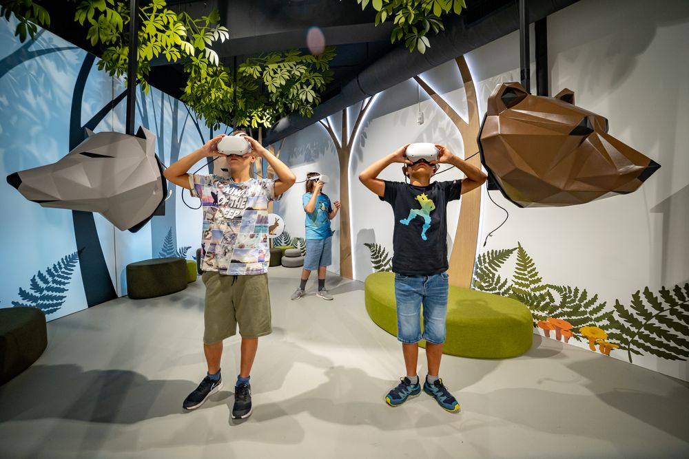 Tre bambini con visori VR esplorano una foresta virtuale in uno spazio espositivo interattivo. Accanto a loro sono sospese sculture stilizzate di un lupo e di un orso, circondate da alberi e decorazioni ispirate al bosco.